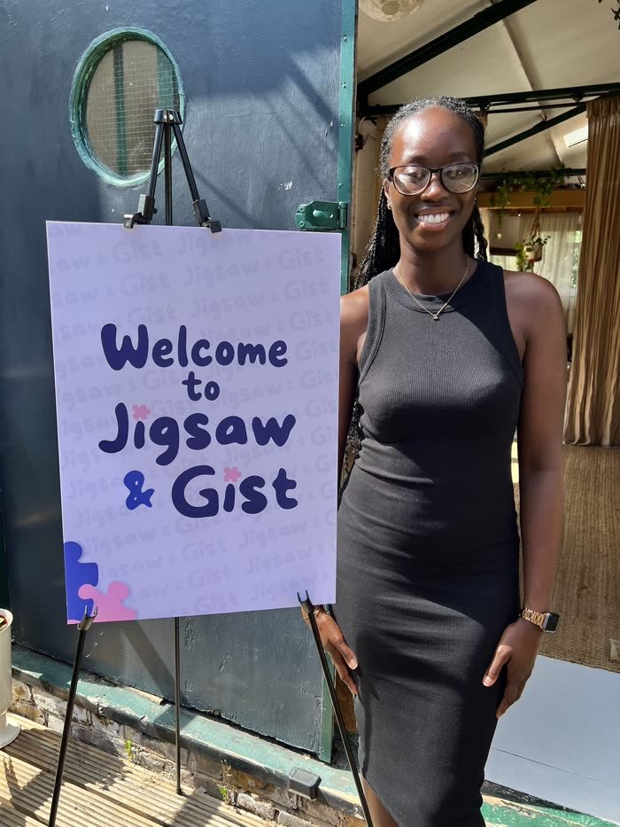 Bethany Ofosuhene, smiling, next to the Welcome to Jigsaw & Gist sign