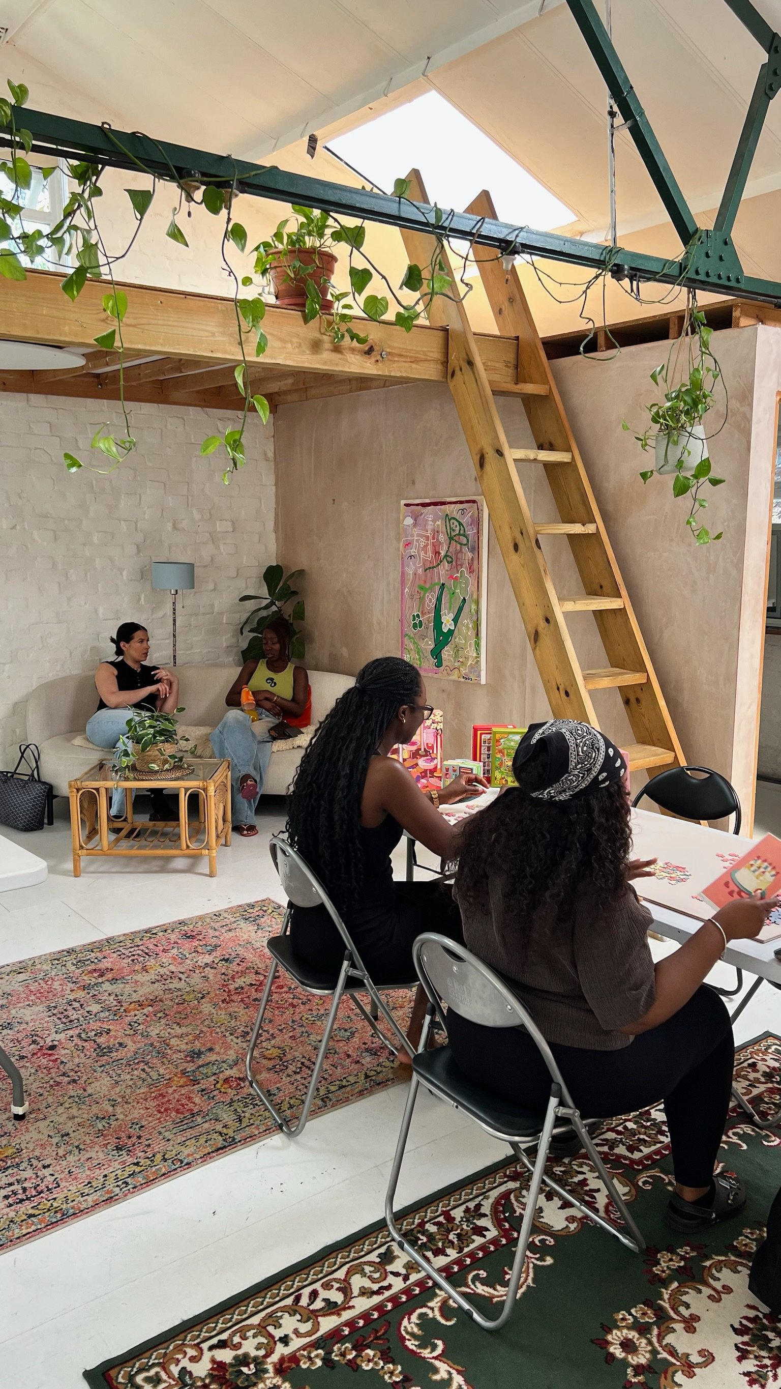 Two people chatting on a sofa while others puzzle at a table nearby