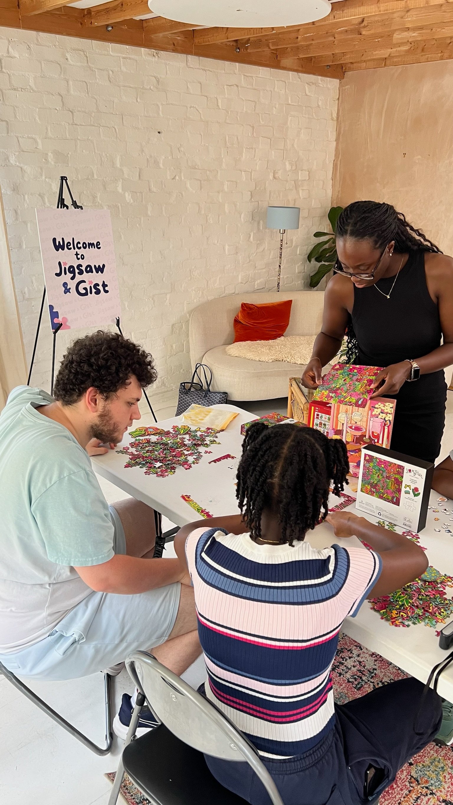 Three people working on a puzzle beside the Welcome to Jigsaw & Gist sign