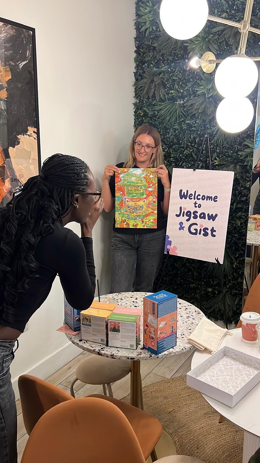 Two people holding up a finished puzzle print beside the Welcome to Jigsaw & Gist sign