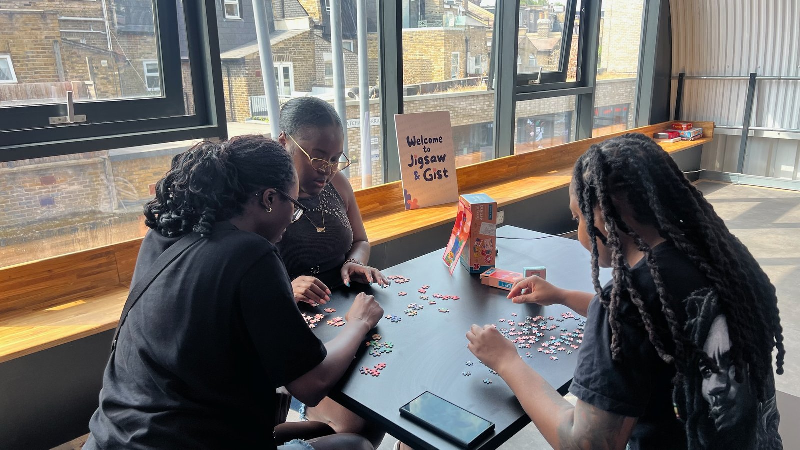 Three people working on a puzzle beside a window, next to the Welcome to Jigsaw & Gist sign